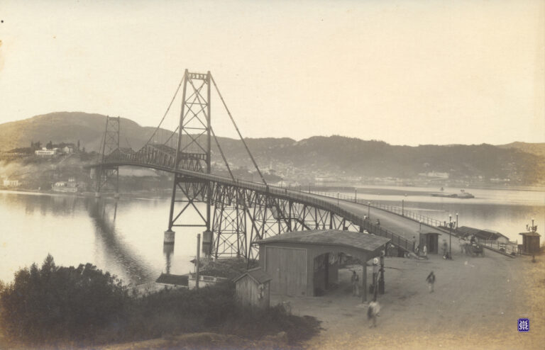 Fotografia histórica em tons de sépia, capturada em plano aberto, apresentando a Ponte Hercílio Luz em Florianópolis.