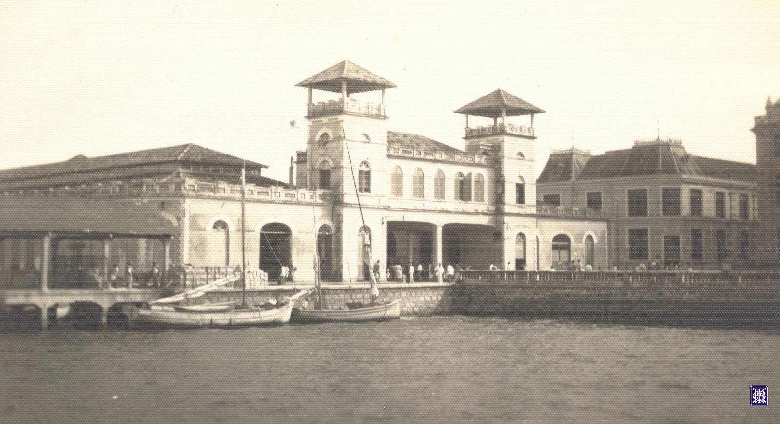 Esta fotografia histórica, em tons de sépia, revela uma movimentada cena portuária. O destaque é a arquitetura imponente do Mercado Público de Florianópolis, ainda à beira-mar
